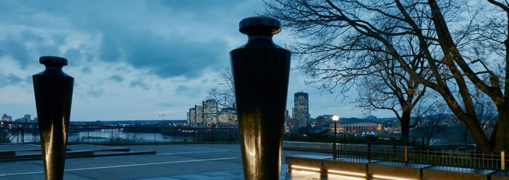 Twilight view of a cityscape, with a pair of oversized plumb bobs, constructed from Cambrian Black granite and mounted on stainless steel pins, illuminated buildings across a river under a cloudy sky.