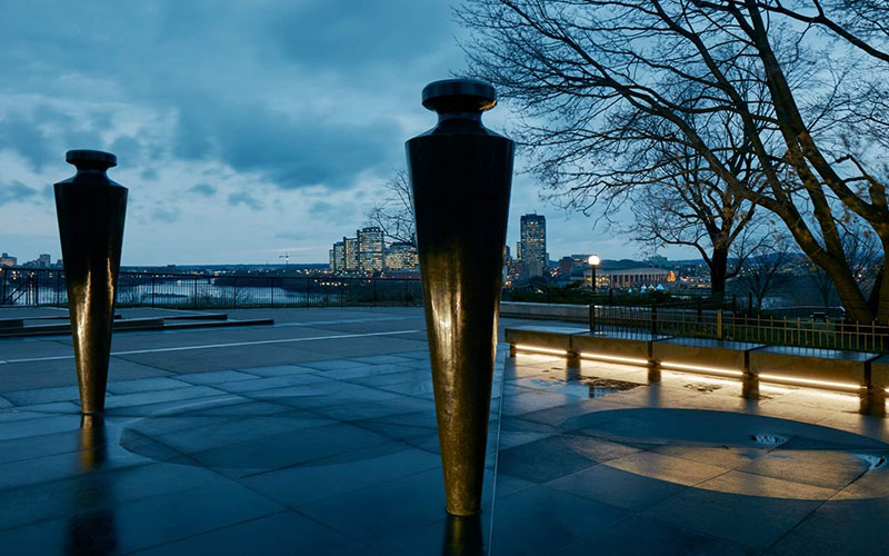 Twilight view of a cityscape, with a pair of oversized plumb bobs, constructed from Cambrian Black granite and mounted on stainless steel pins, illuminated buildings across a river under a cloudy sky.