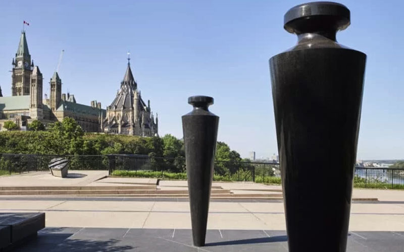 A pair of oversized plumb bobs, constructed from Cambrian Black granite and mounted on stainless steel pins stand on a plaza, with a historic, castle-like building and trees in the background under a clear sky.