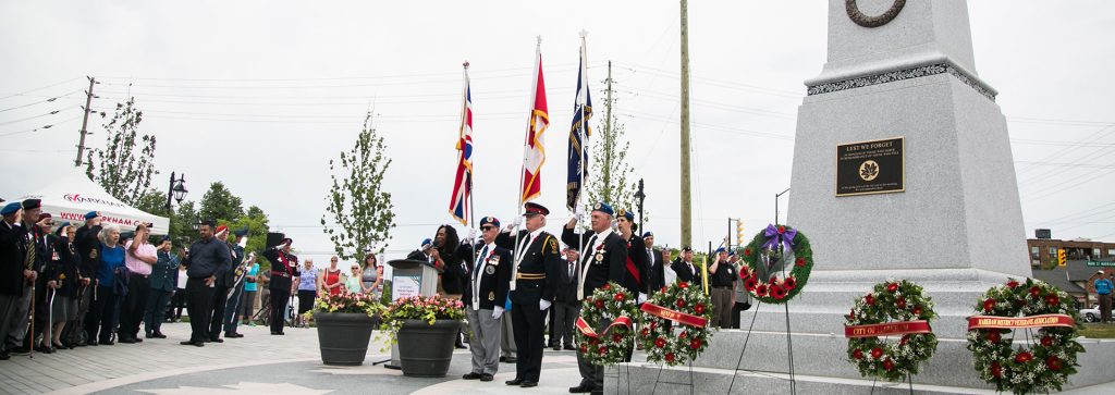 A group of veterans in uniform stand solemnly with flags and wreaths at a memorial ceremony.