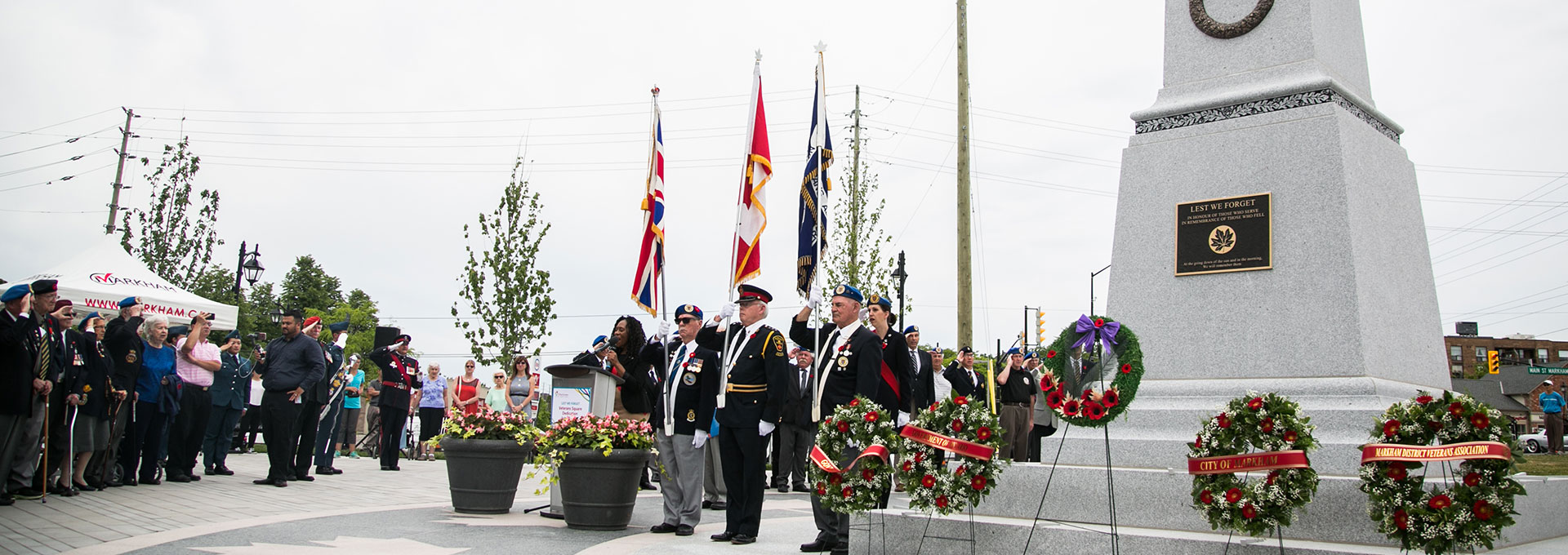 A group of veterans in uniform stand solemnly with flags and wreaths at a memorial ceremony.