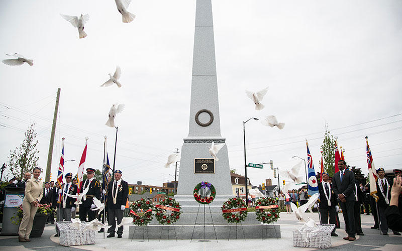 Ceremony at a granite monument with officials, flags, and wreaths. White doves fly upwards, symbolizing peace. The scene is solemn and respectful.