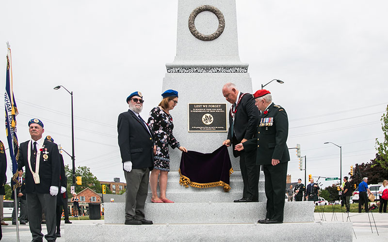 A group of veterans and officials unveils a war memorial plaque with "Lest We Forget" inscribed. The mood is solemn, with flags and military attire.