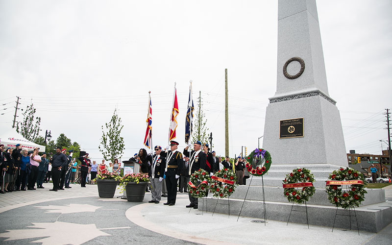 A ceremony at a war memorial with veterans in uniform standing beside flags. Wreaths are placed at the monument's base, surrounded by onlookers.