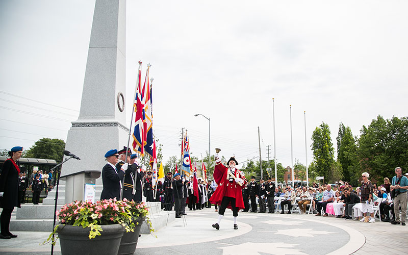 A ceremony at a memorial site, with a town crier in red robes ringing a bell, flag bearers in the background, and a seated audience observing.