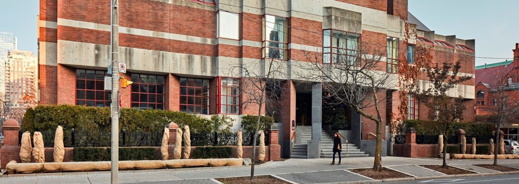 A brick and concrete building with large windows and geometric design sits behind a row of trimmed shrubs and stone sculptures. A person walks nearby.