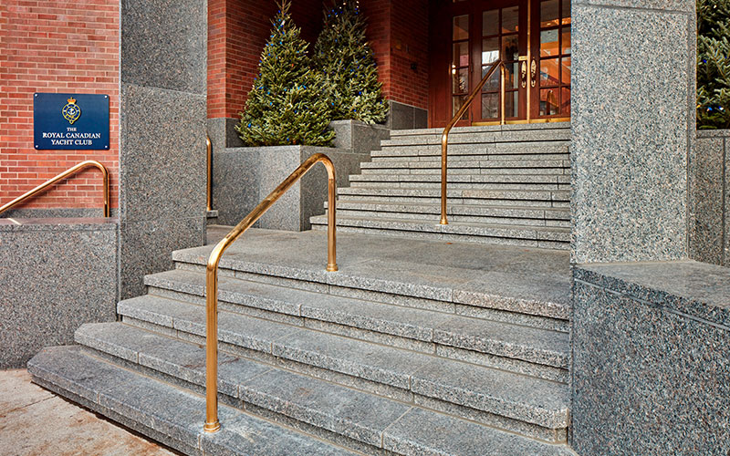 Granite steps with gold handrails lead to a wooden door at the Royal Canadian Yacht Club entrance. Evergreen shrubs flank the stairs, creating an elegant feel.