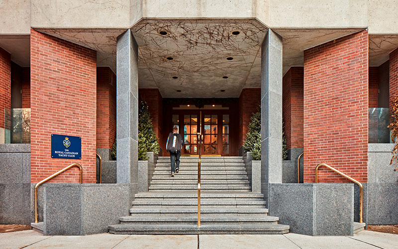 Brick building entrance with wide granite steps and railings, flanked by planters. A person descends the steps. Sign reads "Royal Canadian Yacht Club."