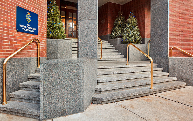 Granite steps with brass railings lead to the entrance of a brick building. A sign reads "The Royal Canadian Yacht Club"; small trees flank the stairs.