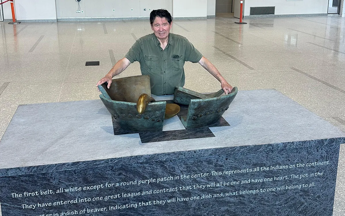 A smiling man in a green shirt poses behind a split bronze sculpture in a bright museum hall, with an engraved inscription in the foreground.