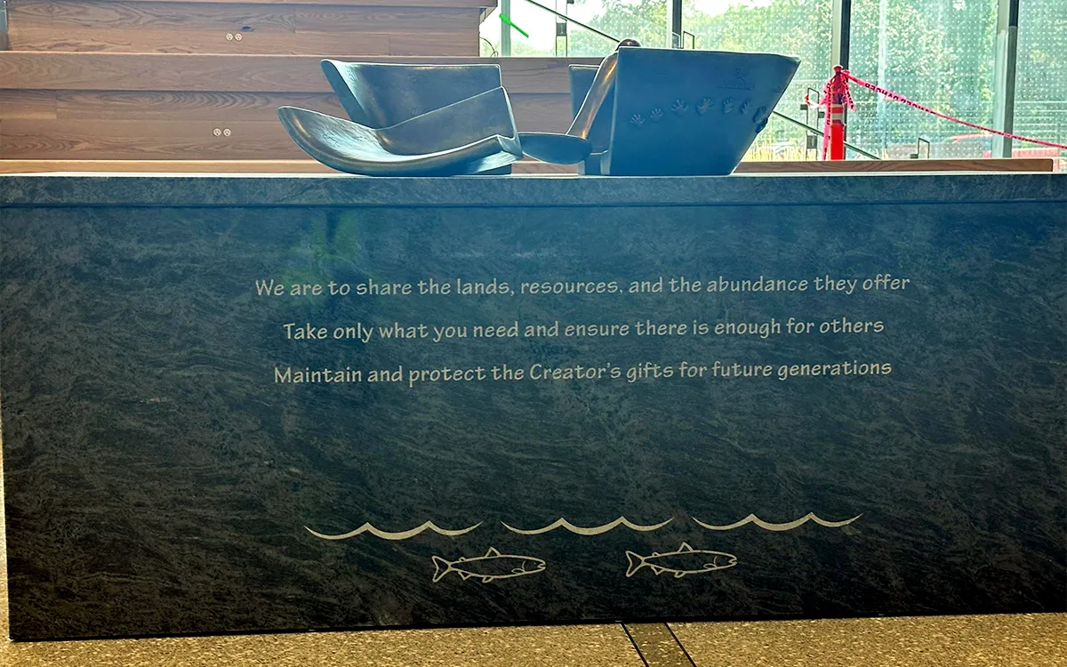 A granite pedestal with a bronze sculpture on top. The front of the pedestal is inscribed with text along with an image of two salmon beneath wavy lines.
