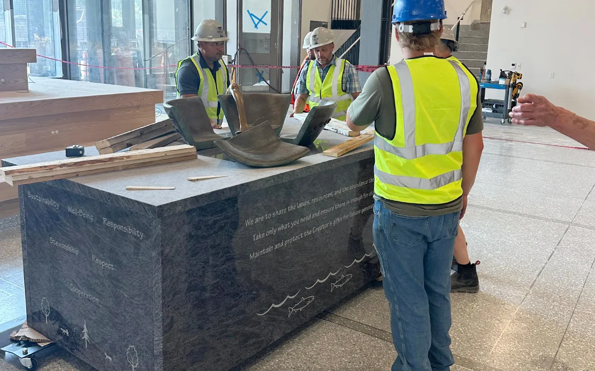 Construction workers in hard hats and reflective vests inspect a curved metal piece on an engraved dark stone monument in a bright workshop.