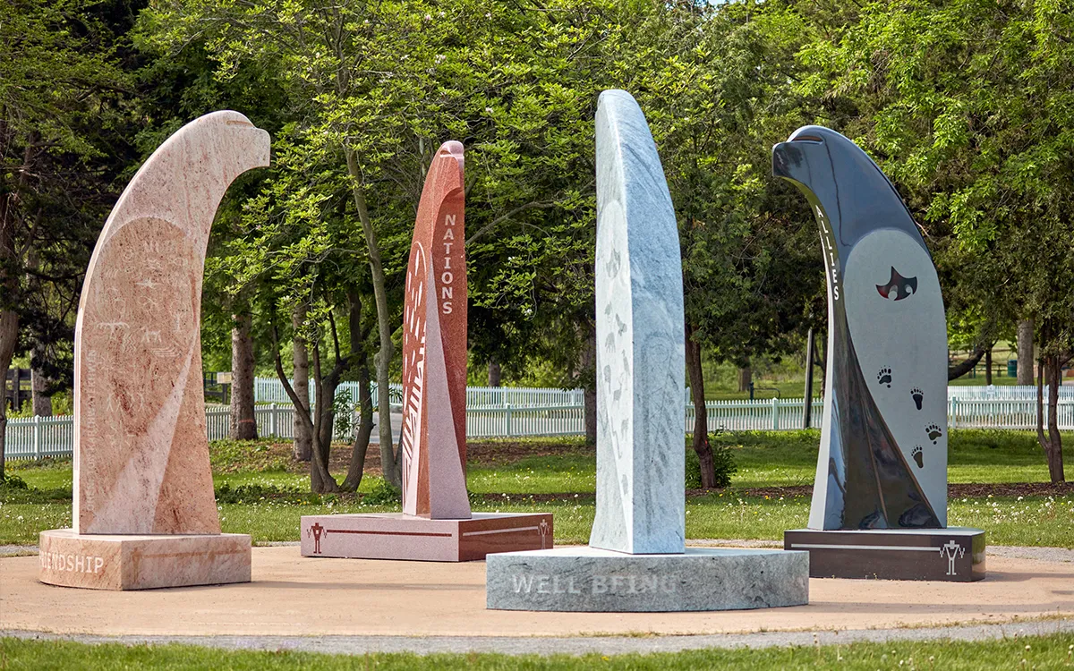Four curved stone sculptures stand in a tree-lined park, engraved with words including “Nations” and “Well Being”; the scene feels peaceful and reflective.