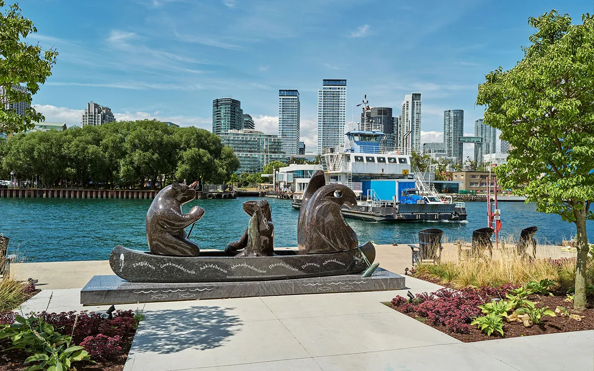 Bronze otter family sculptures sit beside a sunny waterfront, with a boat, landscaped flower beds, leafy trees, and modern city towers beyond.