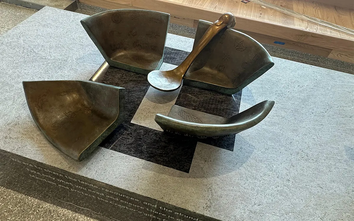 A bronze sculpture of three broken bowl fragments and a spoon on a pedestal with engraved text.